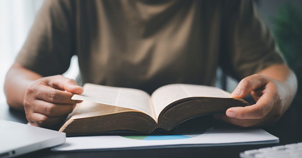 A close-up of hands holding a Bible and reading it at a desk.