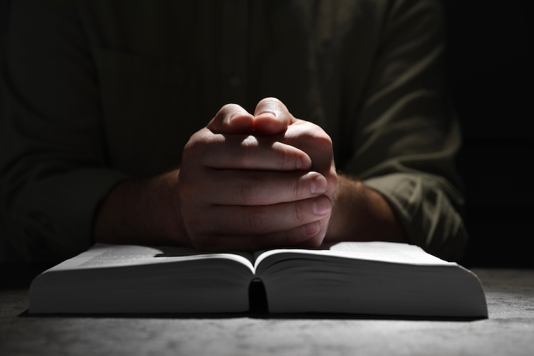 A pair of hands clasped together in prayer over an open bible, evoking the idea of spiritual warfare.