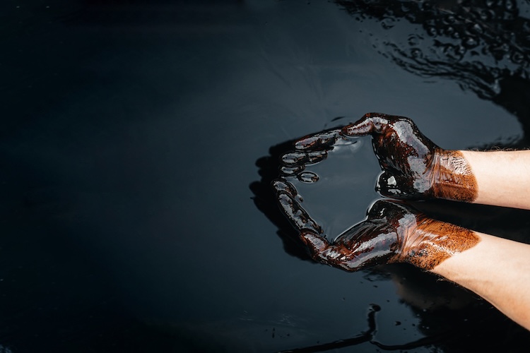 Two hands covered in dark oil cupped together over a black pool of oil.