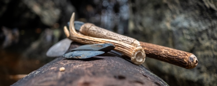Collection of primitive tools with wooden handles and blades resting on a log, representing how archaeology points to creation.