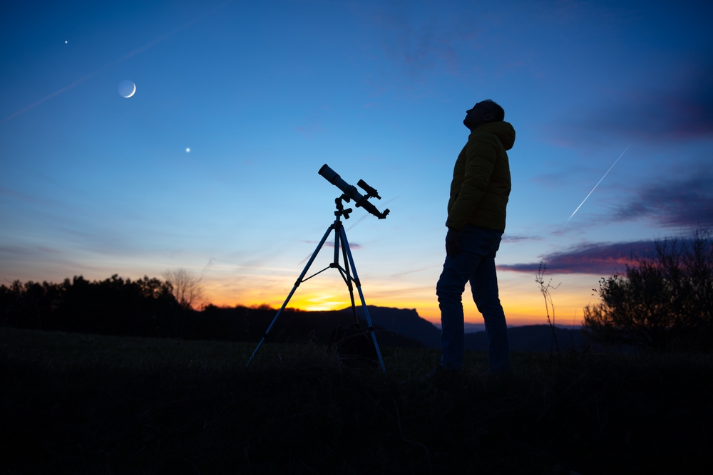 A person stands silhouetted against a vibrant sunset sky with a telescope. A crescent moon and stars are visible in the sky.