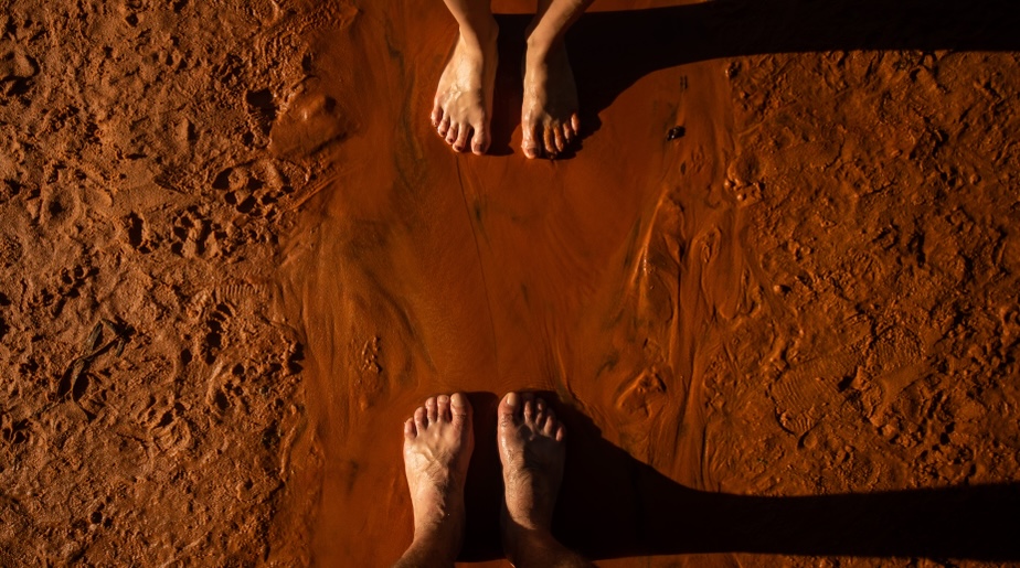 Two pairs of bare feet on textured orange clay earth, representing the sixth day of creation.