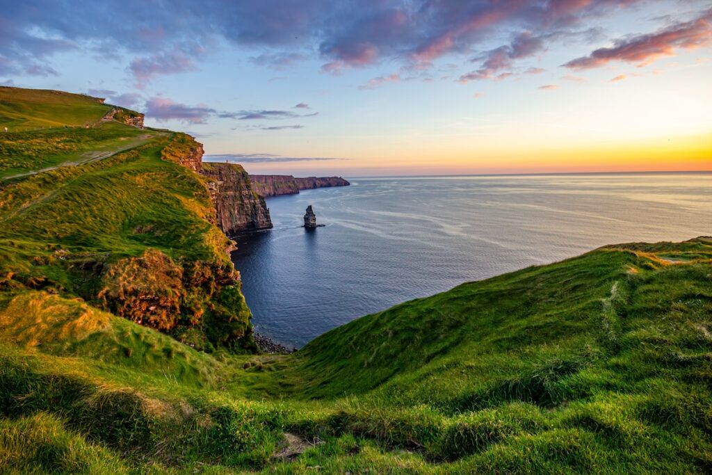 A scenic view of cliffs overlooking a body of water during sunset, with green hills in the foreground and rock formations in the sea, representing the third day of creation.