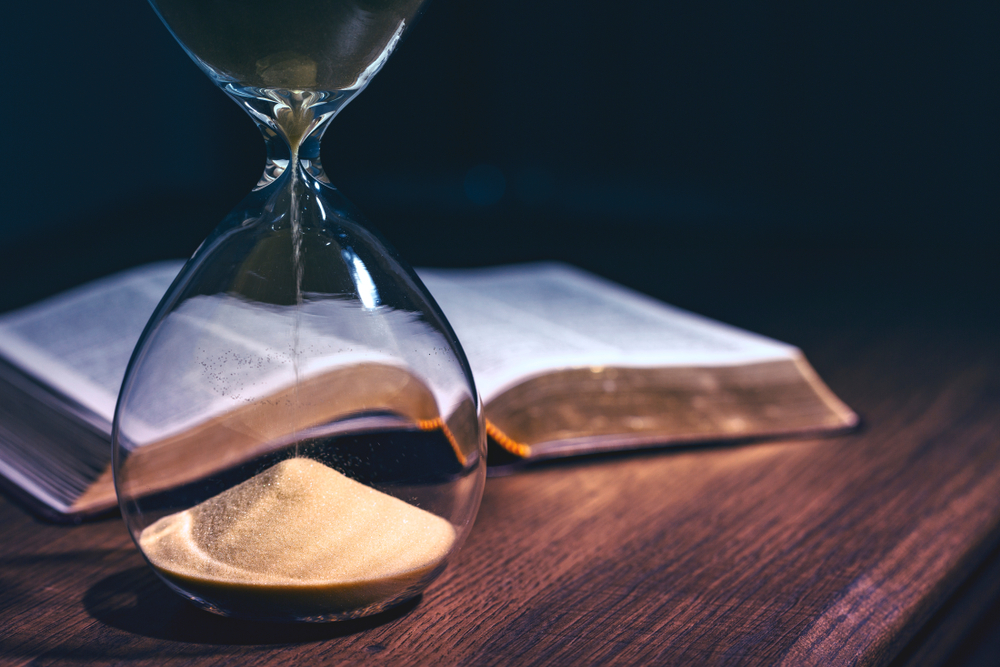 Hourglass resting in front of an open Bible on a wooden table, symbolizing old earth creationism and its interpretation of the “days” of Genesis as long ages rather than literal 24-hour periods.