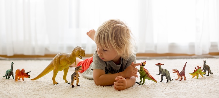 Young child lying on a soft carpet, surrounded by colorful dinosaur toys.