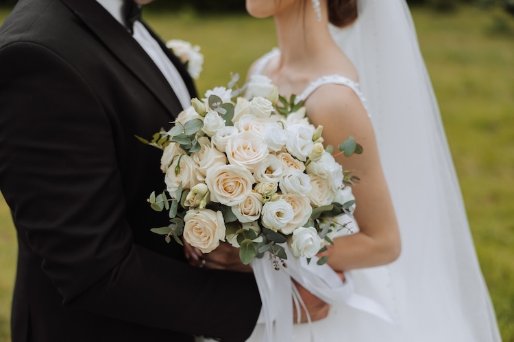 A bride and groom standing outdoors, shown from the shoulders down, with the bride holding a bouquet of ivory and white roses accented with greenery, symbolizing the beauty and dignity of human beings made in the image of God, male and female.