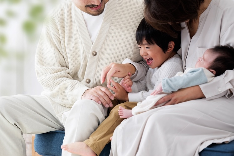 A father and mother sit together on a sofa, gently holding and supporting two young children between them, illustrating human beings made in the image of God through loving, attentive care.