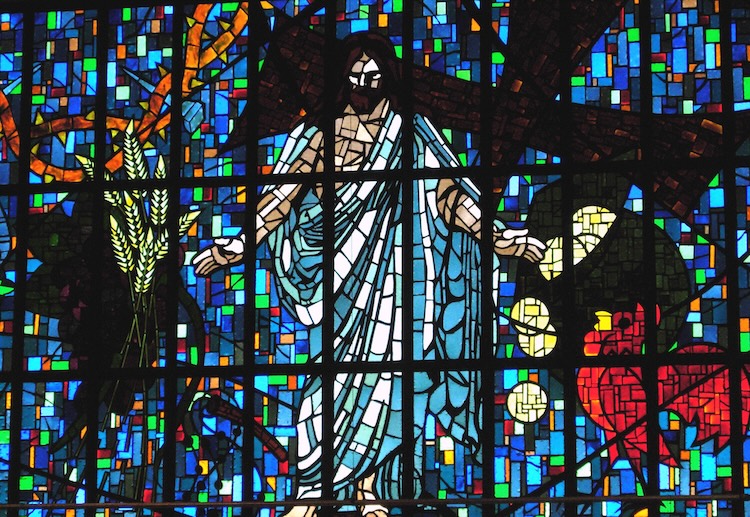 Stained-glass artwork depicting Jesus—the perfect image of God the Father—with outstretched arms in front of a cross, surrounded by wheat, a rooster, and a bag of gold coins.