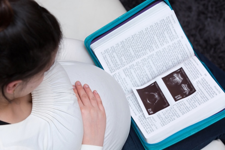 A pregant woman resting her hand on her abdomen while reading an open Bible with two ultrasound images resting on the pages.