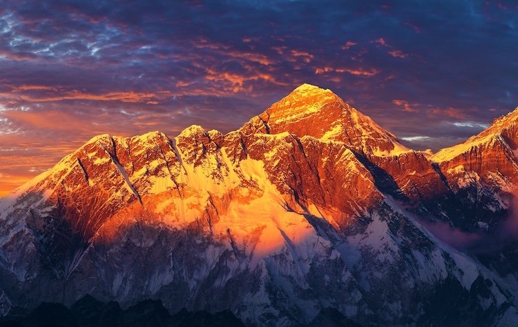 Majestic snow-capped mountains bathed in warm sunset hues and deep shadows under a dramatic sky with scattered clouds.