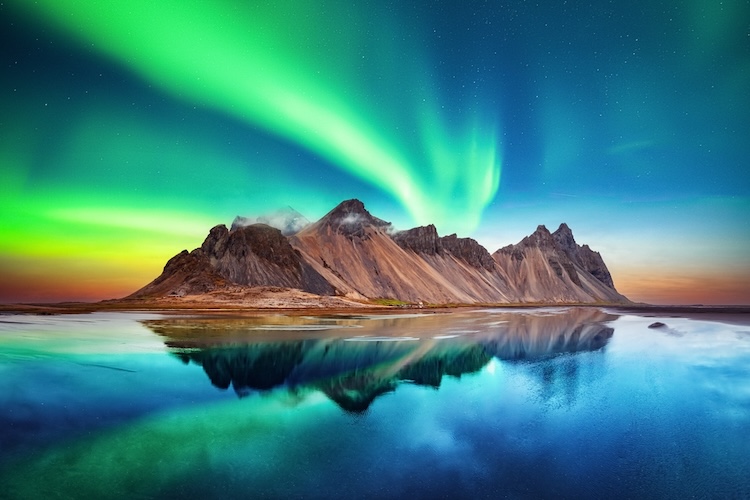 Northern lights glow vibrantly over jagged mountains, mirrored in a tranquil lake below.