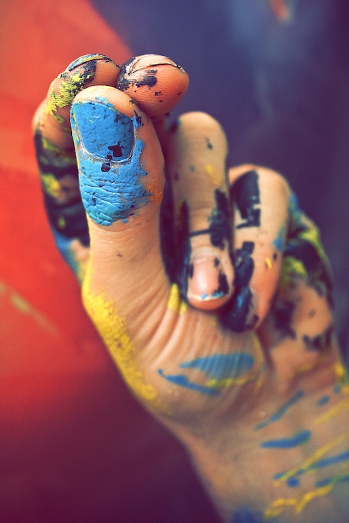 Close-up of a hand covered in vibrant blue, yellow, and green paint against a blurred red and blue background.