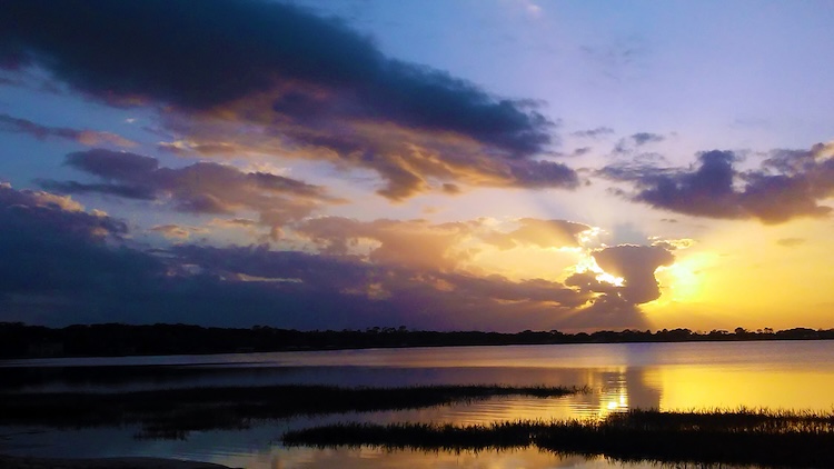A serene sunset over a calm lake, with vibrant purple and orange hues in the sky. Dark clouds partially obscure the sun, reflecting on the water