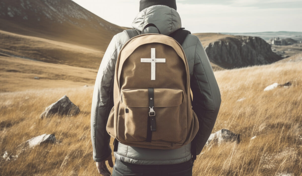 A person wearing a backpack with a cross symbol on it, walking across a grassy landscape with mountains in the background.