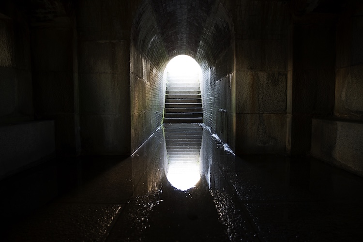 Stairs rise from a flooded dark tunnel toward bright light, symbolizing hope and salvation.