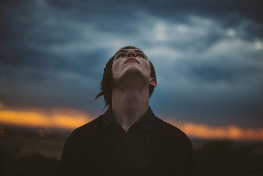 Person gazes upward against a dramatic sky with dark clouds and a warm orange sunset glowing along the horizon.