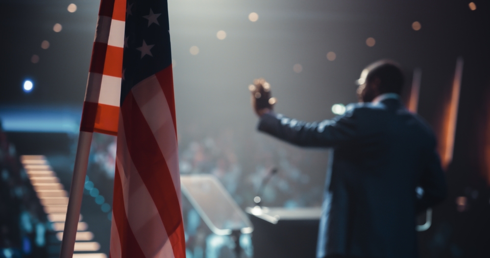 Speaker addressing a crowd beside an American flag, illustrating debates around Christian nationalism.