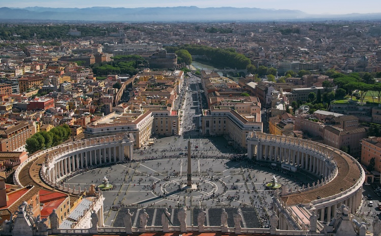 An aerial view of St. Peter’s Square and the surrounding architecture of Vatican City shows the large oval colonnade, central obelisk, and broad avenue leading into Rome, representing one of the historic centers of Christianity and the several splits within Christianity over the centuries.