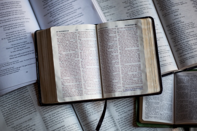 An open Bible with highlighted passages rests on top of several other open Bibles, representing the central role of Scripture in Christian beliefs and how God reveals himself through His Word.