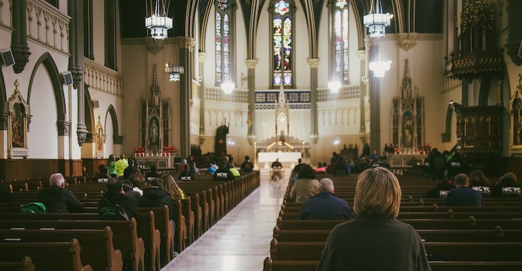 The interior of a large, ornate church shows rows of wooden pews leading toward a bright altar, with stained‑glass windows, tall arches, and detailed architectural features that reflect historic Christian traditions and places of worship.