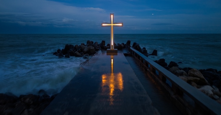 A cross—the most common symbol of Christianity—stands illuminated by glowing lights at the end of a wet stone pier extending into the ocean at dusk.