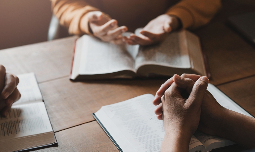 Several open Bibles rest on a wooden table while a small group sits in discussion, illustrating the study of Scripture that is central to Christian beliefs and practices.