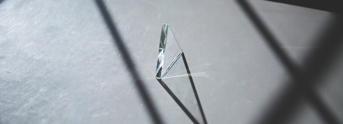 A small triangular glass prism standing on a surface in sunlight, casting a long shadow.