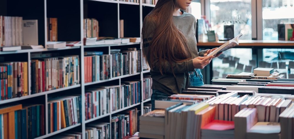 A young woman browses books in a bookstore aisle, holding one open as she looks through it, surrounded by shelves filled with many titles—representing a thoughtful search for truth, meaning, and understanding among different worldviews.