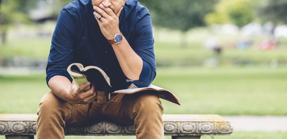 A man in a blue shirt sits on a stone bench in a park, thoughtfully reading a book. He rests his chin on hand, conveying contemplation.