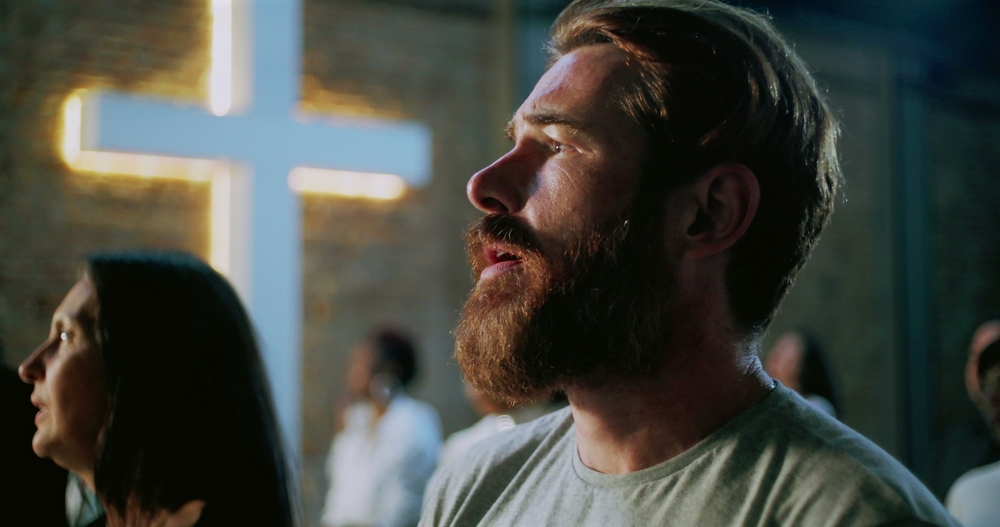 A man with a beard looks upward with a contemplative expression in a church. A glowing cross is visible on the brick wall, and others are in the background.