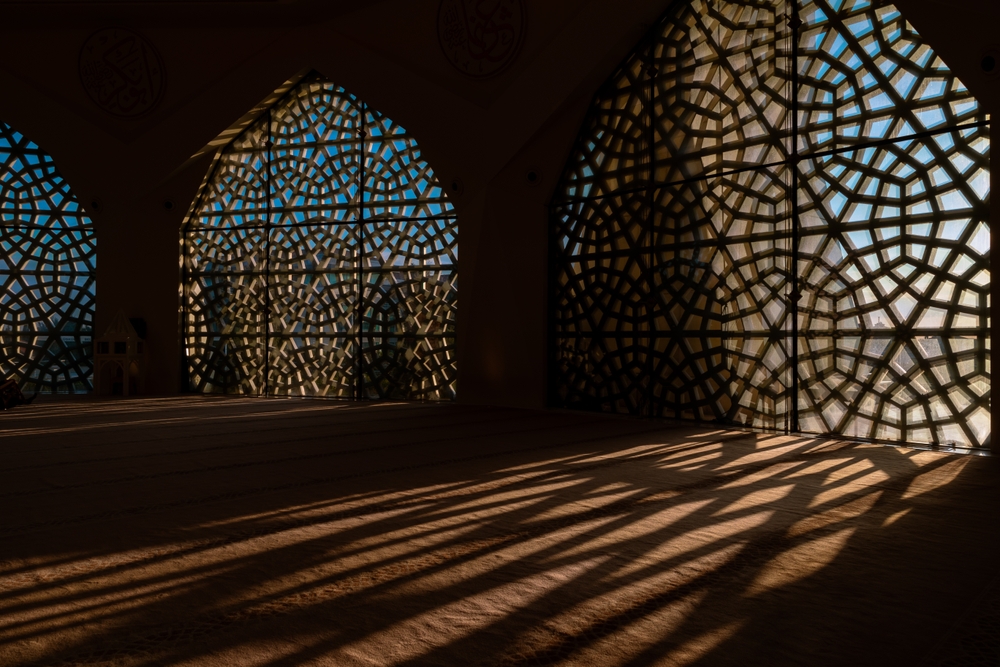 Ornate lattice windows cast intricate shadows on a sunlit floor in a mosque interior.
