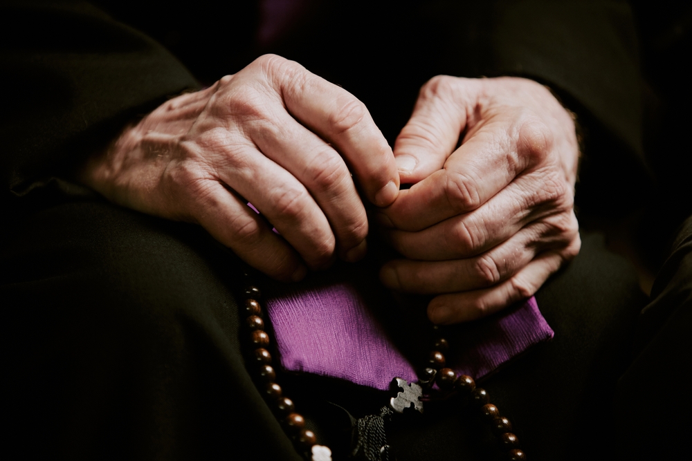 Close-up of an older person’s hands resting in their lap, holding a rosary over a small purple cloth.