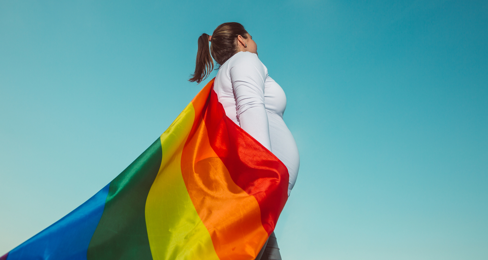Pregnant person standing against a blue sky wearing a rainbow Pride flag draped like a cape