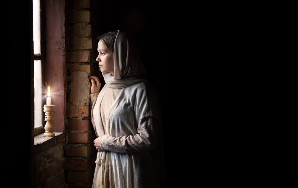 A contemplative woman beside a candle looking out a window, illustrating themes of faith and the historical role of women in Christianity.