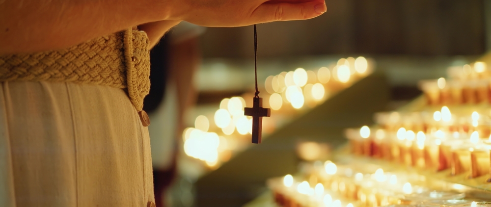 A closeup of a woman's hand holding a cross, a symbol of Christian faith, beside rows of lit candles.
