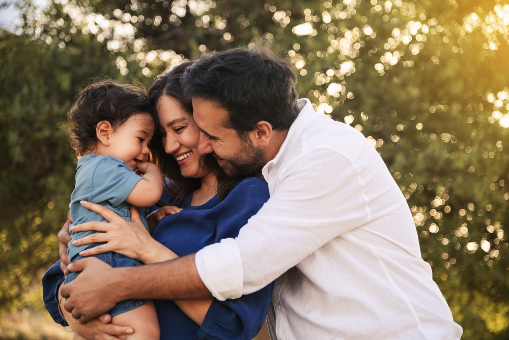 A father and mother hold their toddler with trees in the background and the sun shining through the trees.