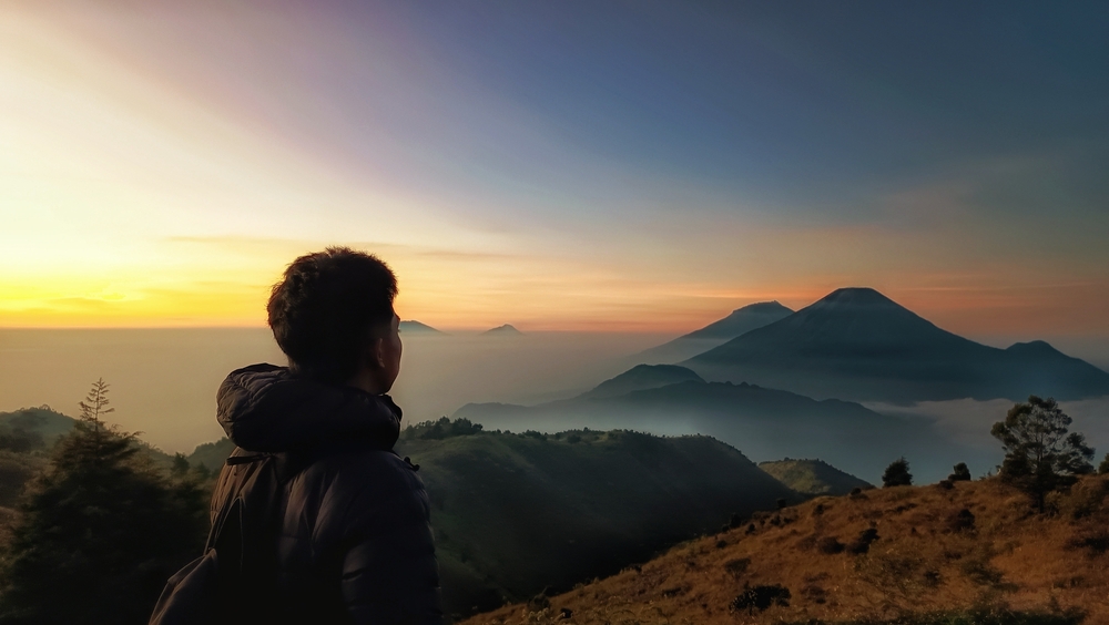 A young man with a backpack surveys the mountains ahead of him, representing the highs and lows in the lives of Christians who trust in God's faithfulness.